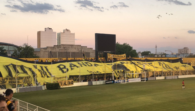 Ein Fußballfeld mit einer Gruppe von Menschen darauf, umgeben von einem Metallzaun, einem Torpfosten, einer Fahne mit der Aufschrift "Boca Juniors vs Real Sociedad", einigen Pflanzen, einem Anzeigebildschirm, einigen Pfosten, der Rinde eines Baumes, einigen Bäumen, einigen Gebäuden und einem bewölkten Himmel.