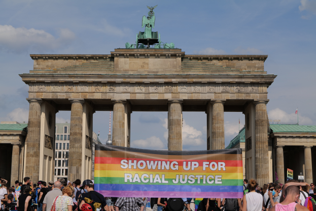 Eine Gruppe von Menschen steht vor dem Brandenburger Tor in Berlin, Deutschland, und hält eine Fahne mit der Aufschrift "Rassengerechtigkeit". Das Tor hat Säulen und eine Statue, im Hintergrund sind Gebäude und ein bewölkter Himmel zu sehen.