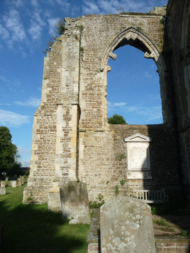 Die Ruinen der St. Mary's Church in St. Margaret's, Suffolk, England, mit einem Bogen, umgeben von Gras, Grabsteinen, Bäumen und einem bewölkten Himmel.