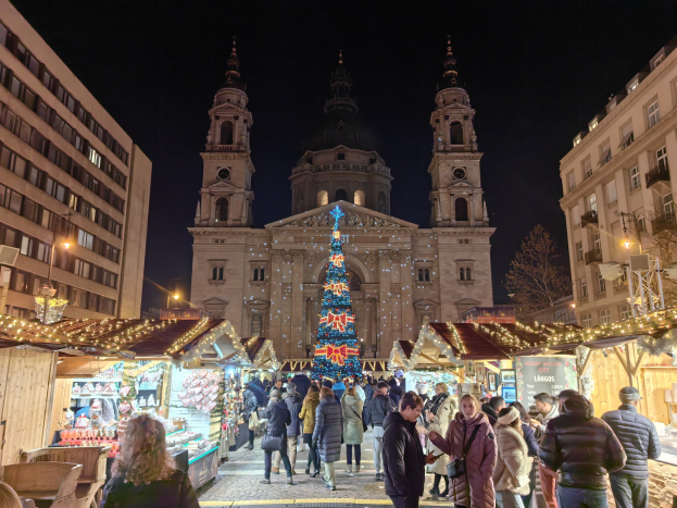Ein belebter Weihnachtsmarkt mit geschmückten Ständen und Menschen vor einer Kirche bei Nacht, vor Buildings, Bäumen und einem sternenklaren Himmel.