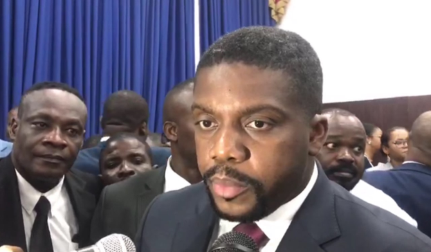 Ein Mann im Anzug und Krawatte spricht vor einer Gruppe von Menschen in ein Mikrofon, mit Vorhängen und einer Wand im Hintergrund, und scheint auf eine lebenslange Haftstrafe zu reagieren.