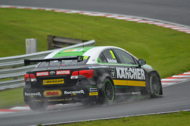 Ein Auto mit Text an der Seite fährt auf einer regennassen Rennstrecke, die von Gras gesäumt ist, mit einer Begrenzung und einem hölzernen Aufbau im Hintergrund.