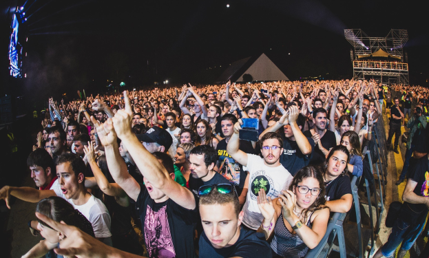 Menschenmenge auf einem Musikfestival mit erhobenen Händen, sitzend und stehend, hinter einem Metallzaun, mit Gebäuden und Lichtern im Hintergrund unter einem dunklen Himmel.