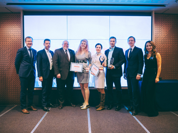 Gruppe von strahlenden Menschen, die Zertifikate in die Kamera halten, vor einem Bildschirm und einem Tisch mit Wandobjekt im Hintergrund, Gewinner der Internationalen Business Awards 2018.