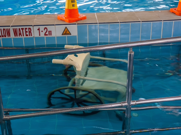 Ein Schwimmbad mit einem Stuhl in der Mitte, umgeben von Sicherheitskegeln und einem Geländer, sowie einem Schild an der Wand, das die Anwesenheit von Schwimmausrüstung anzeigt.
