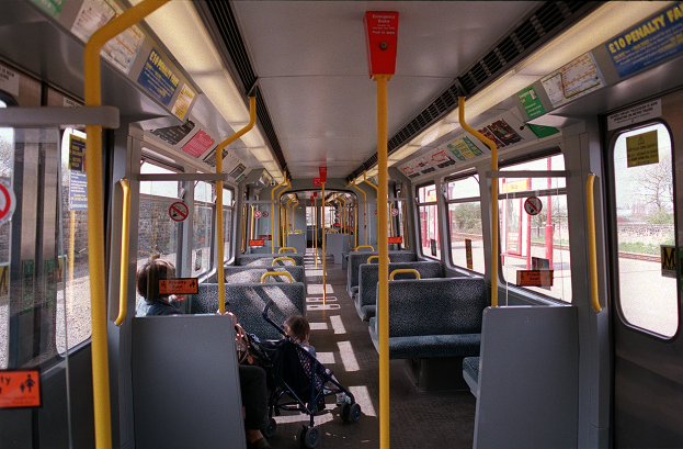 Das Innere eines Zugwagens mit Menschen auf den Sitzen, einem Baby im Kinderwagen, offenen Fenstern und Schildern mit Text.