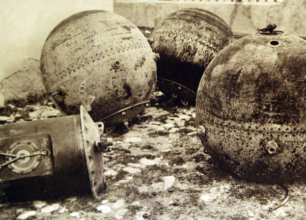 Drei große Metallbomben stehen auf dem Boden vor einer Wand mit einem Fenster im Hintergrund, dargestellt in einem Schwarz-Weiß-Foto.