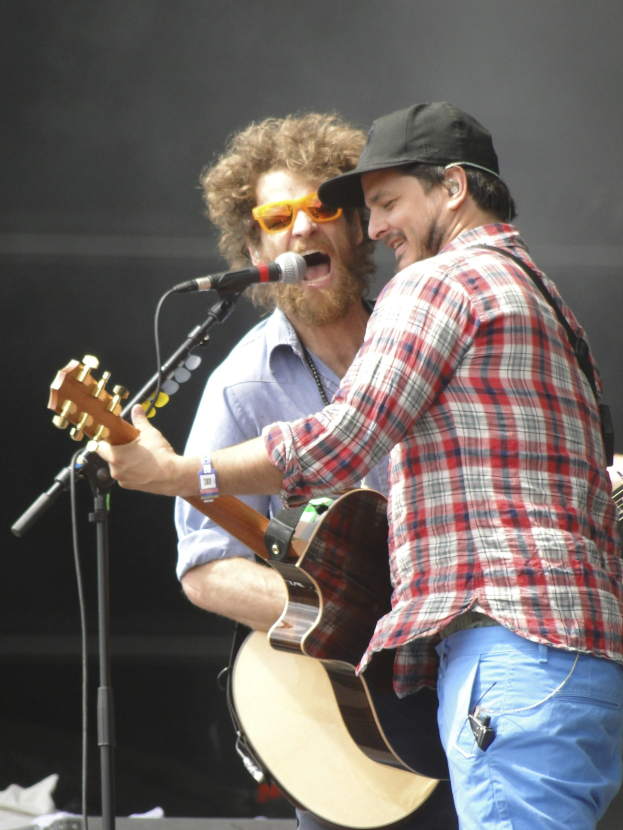 Zwei Männer auf der Bühne mit Gitarren und Mikrofonen, einer in rot-blau kariertem Hemd und blauen Hosen, der andere mit Brille und schwarzer Mütze.