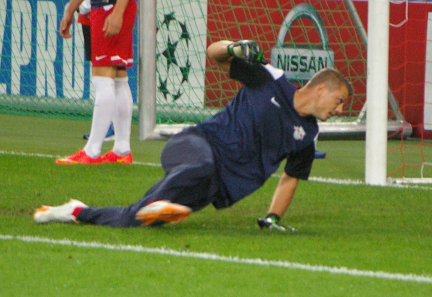 Ein Fußballspieler in Handschuhen taucht ab, um einen Ball vor einem Tor zu fangen, mit zwei Personen, einem Netz und einer Banner mit Text im Hintergrund.