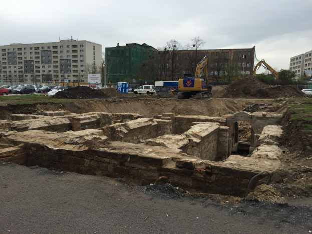Baustelle mit einem großen Haufen Erde, umgeben von Baggern, Fahrzeugen, Gras, Bäumen und Gebäuden mit Fenstern im Hintergrund unter einem bewölktem Himmel, mit einer Straße unten.