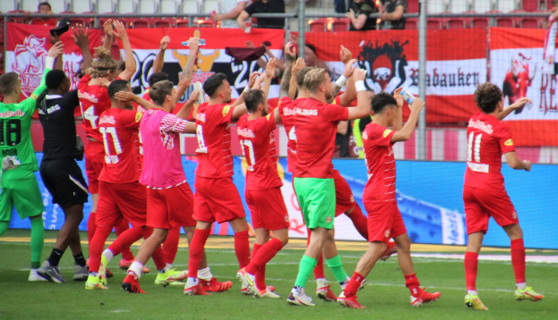 Gruppe von Fußballspielern, die ein Tor feiern, auf einem Feld, einige halten Flaschen oder klatschen, mit einem Zaun, Bannern, Metallstangen, Stühlen und einem Dach mit Deckenleuchten im Hintergrund.