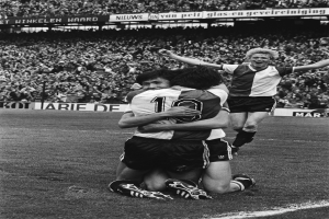 Drei Fußballspieler umarmen sich auf dem Boden, um ein Tor zu feiern, mit einer jubelnden Menge und schwenkenden Fahnen im Hintergrund, dargestellt in Schwarz-Weiß.