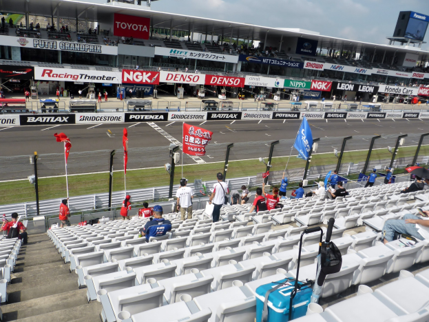 Gruppe von Menschen auf Tribüne bei einer Rennstrecke mit Fahnen, Texttafeln, Geländern und Stufen, mit bewölktem Himmel im Hintergrund.