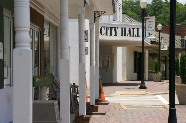 Ein Rathaus umgeben von Gebäuden, Straßenlaternen, Namensschildern, Kraftfahrzeugen, Verkehrskegeln, Stühlen, Topfpflanzen, Bäumen und einem bewölkten Himmel.