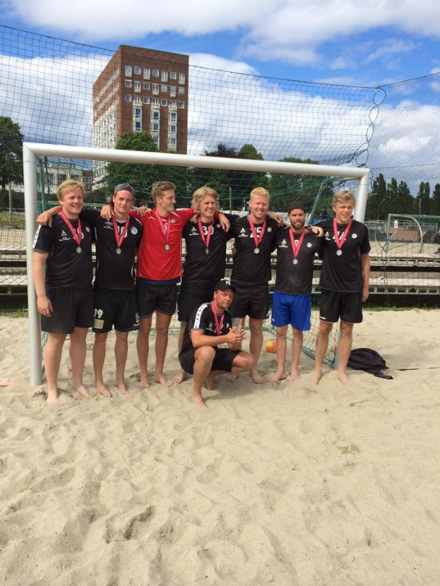 Mannergruppe in Sportkleidung auf einem sandigen Strand, mit Medaillen und umgeben von einem Netz, mit Bäumen, Gebäuden und einem bewölkten Himmel im Hintergrund.