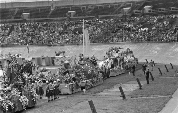 Schwarzes und weißes Foto einer Parade in einem Stadion mit Menschen, die stehen und sitzen, einem zentralen Springbrunnen und Blumenbouquets auf Fahrzeugen.
