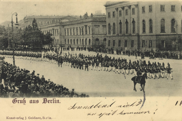 Ein altes Schwarz-Weiß-Foto von einer Parade in Berlin, Deutschland, mit Menschen auf dem Boden und auf Pferden, einer Statue auf einem Sockel, Straßenlaternen, Bäumen, Gebäuden mit Fenstern und einem bewölkten Himmel, mit Text am unteren Rand des Bildes.