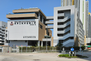 Ein große Gebüude mit zahlreichen Fenstern, identifiziert als Teng Hin Pei Grundschule, umgeben von Straßenmasten, Straßenlaternen, Schildern, Kraftfahrzeugen, Fussgöngern, Bürsten, Böumen und unter einem klaren blauen Himmel.