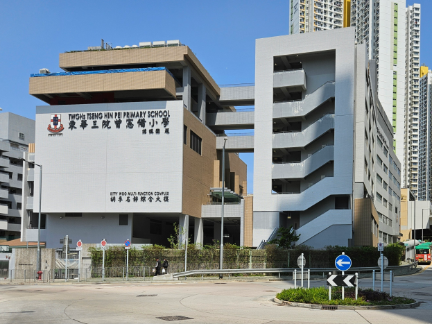 Ein große Gebüude mit zahlreichen Fenstern, identifiziert als Teng Hin Pei Grundschule, umgeben von Straßenmasten, Straßenlaternen, Schildern, Kraftfahrzeugen, Fussgöngern, Bürsten, Böumen und unter einem klaren blauen Himmel.