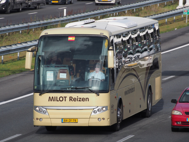Ein Minibus mit Passagieren auf einer Autobahn neben einem roten Auto, mit Geländern und Gras an den Seiten und Texttafeln und Säulen im Hintergrund.