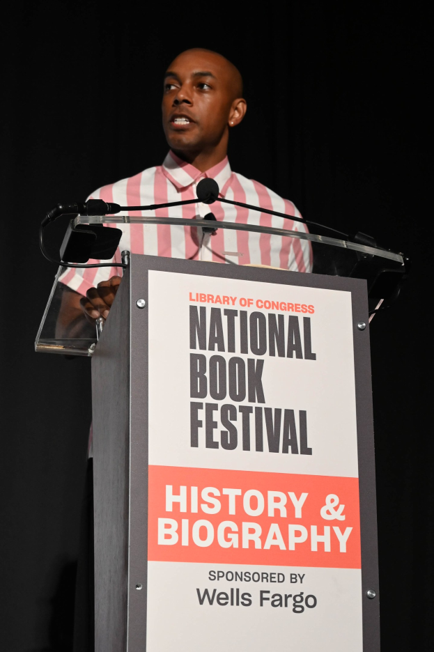 Ein Mann steht an einem Rednerpult mit einem Mikrofon, spricht bei einer Abendveranstaltung mit einem "National Book Festival History & Biography"-Schild dahinter.