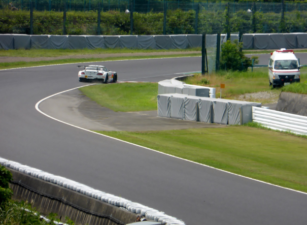 Ein Rennauto, das auf einer Rennstrecke fährt, mit einem Polizeiwagen im Hintergrund, umgeben von Gras, Pflanzen, Bäumen und einem Zaun, mit einem Fahrzeug auf der rechten Seite und Text unten.