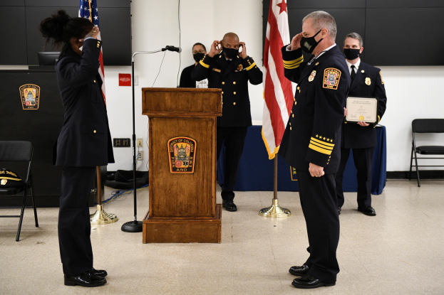 Eine Gruppe uniformierter Personen in Masken steht vor einem Podium mit Mikrofon, umgeben von Fahnen, Stühlen und Tischen, während eine feierliche Vereidigung für einen neuen Feuerchef stattfindet, wobei eine Person ein Zertifikat hält.