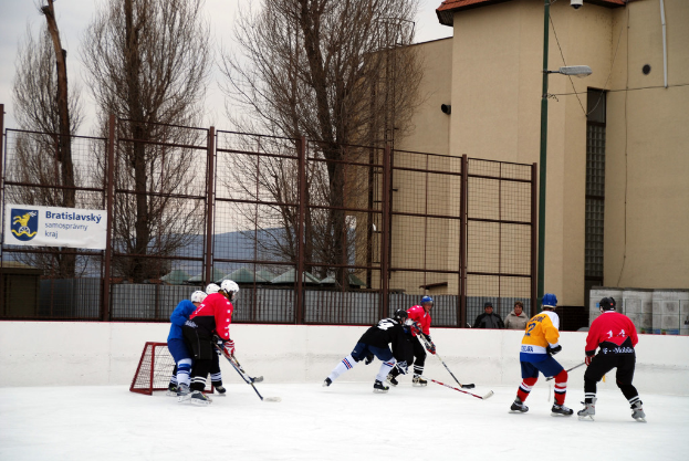Menschen, die Eis hockey auf einer Eisbahn mit Gebäuden, Bäumen, einer Straßenlaterne, einem Namensschild und Zäunen im Hintergrund unter einem klaren Himmel spielen.