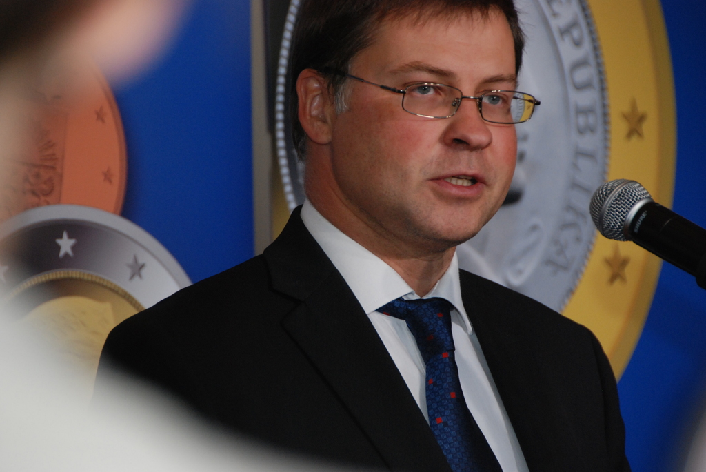 Ein Mann in einem schwarzen Jackett, weißem Hemd und blauer Krawatte spricht in ein Mikrofon vor einem blauen Plakat mit Logos.