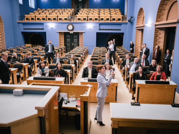 Frau hält Rede im schottischen Parlament, steht vor sitzenden und stehenden Zuhörern mit Tischen, auf denen Mikrofone, Gläser und Papiere stehen, unter Deckenleuchten mit einer Uhr an der Wand dahinter.