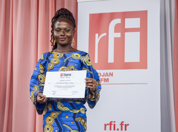 Frau in blauem und gelbem Kleid, die einen Award hält und strahlt, mit einem Banner und Vorhang im Hintergrund.