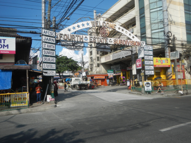 Eine belebte Stadtstraße mit fahrenden Fahrzeugen und Fußgängern, mit einem zentralen Schild, das 'Vierte Gewalt - Unterteilung' bezeichnet, umgeben von Gebäuden, Bäumen, Strommasten und einem bewölkten Himmel.