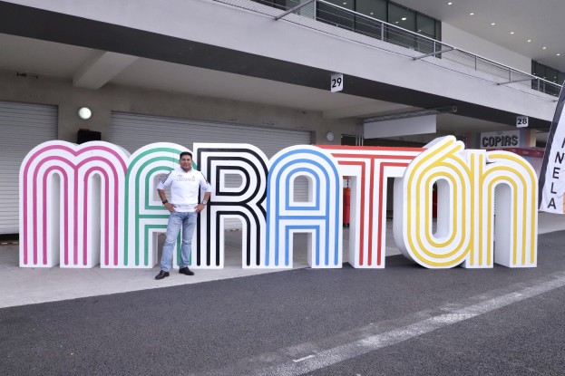 Ein Mann steht am Start eines Marathons vor einem großen "Marathon"-Schild, mit einem Banner rechts daneben und einem Gebäude im Hintergrund.