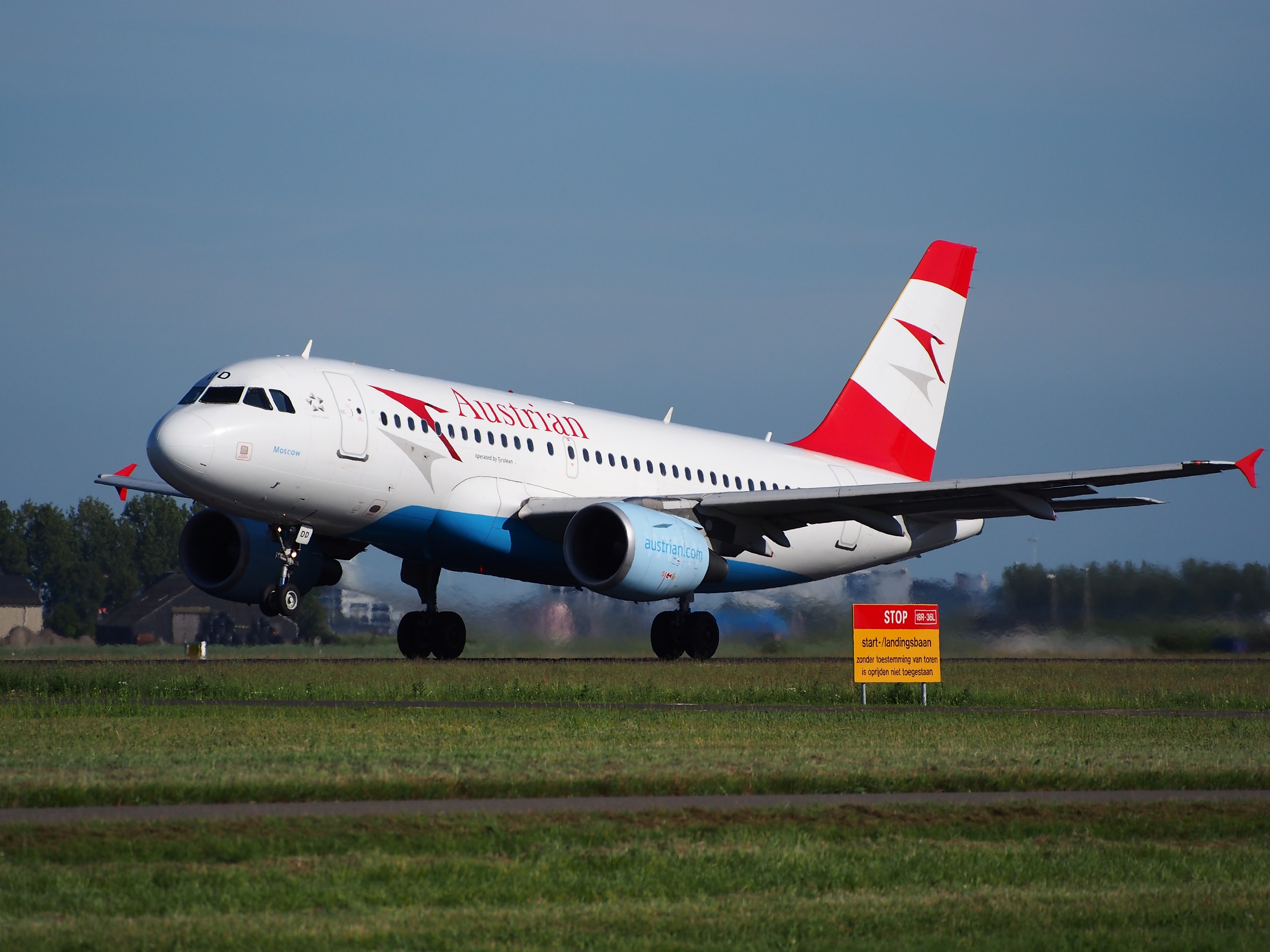 Ein Airbus A320-200 der Austrian Airlines startet vom Frankfurter Flughafen, mit einer Texttafel im Vordergrund, Gras, Bäumen, Gebäuden und einem klaren blauen Himmel im Hintergrund.