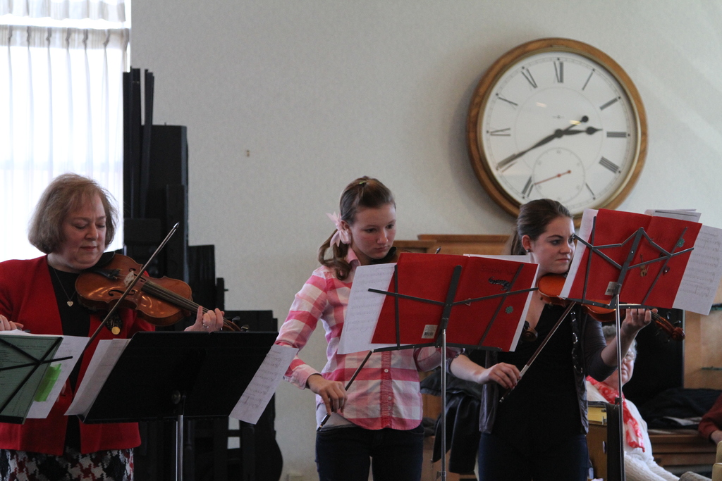 Drei Frauen, die auf der Bühne Geigen spielen, mit Notenpulten und Noten davor, während ein Zuschauer in der foreground sitzt; eine Uhr hängt an der weißen Wand im Hintergrund, und ein weißer Vorhang ist links sichtbar.
