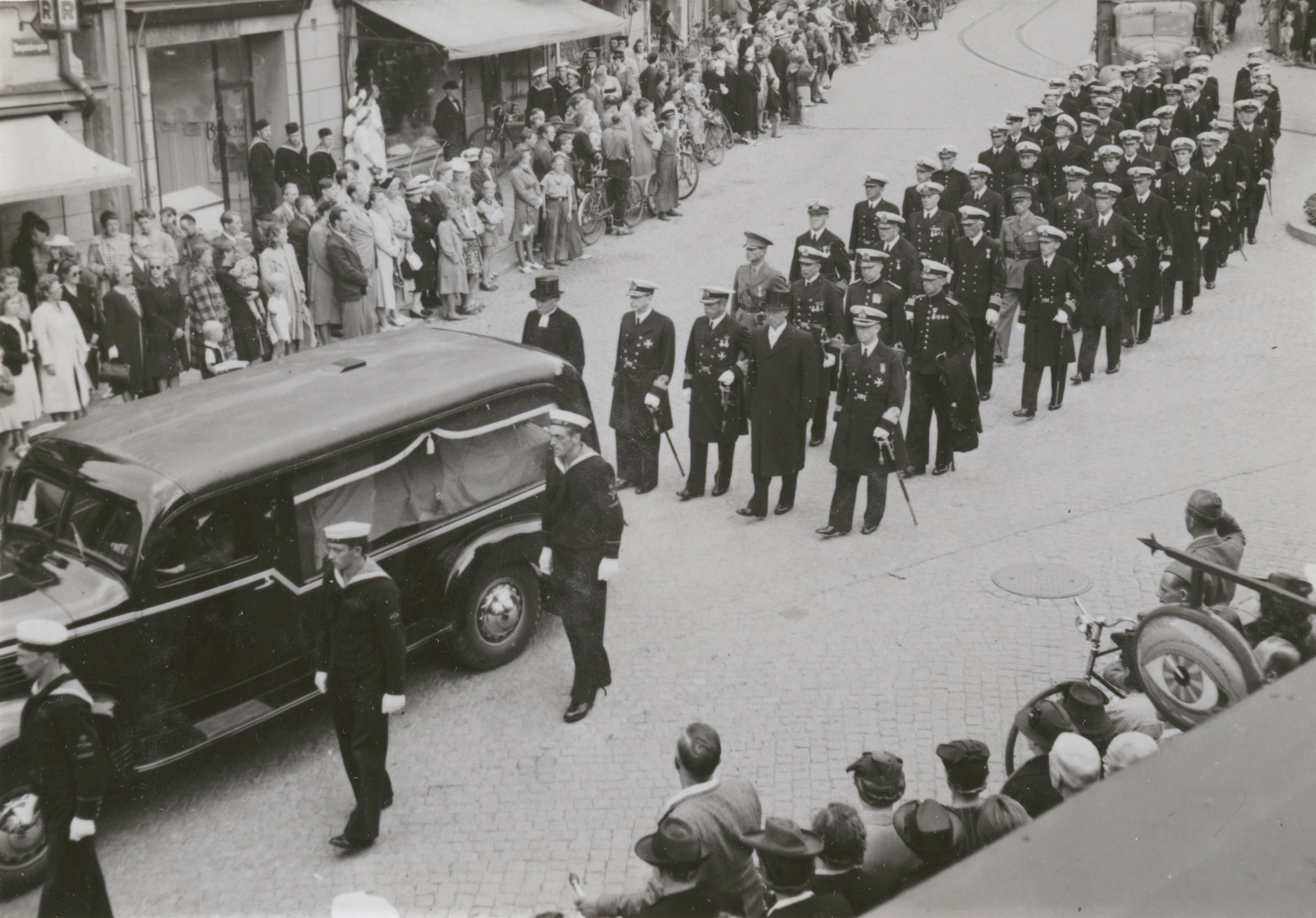 Schwarzes und weißes Bild einer Beerdigungsprozession mit einem Leichenwagen, der den Körper eines Mannes trägt, umgeben von Menschen, einige halten Waffen, Fahrzeuge auf der Straße und Gebäude mit Fenstern und Namensschildern im Hintergrund.