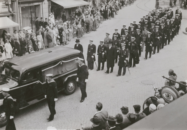 Schwarzes und weißes Bild einer Beerdigungsprozession mit einem Leichenwagen, der den Körper eines Mannes trägt, umgeben von Menschen, einige halten Waffen, Fahrzeuge auf der Straße und Gebäude mit Fenstern und Namensschildern im Hintergrund.