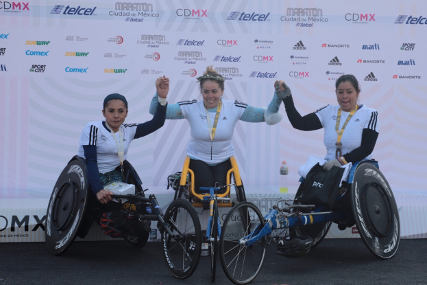Drei Frauen im Rollstuhl auf einem Podium, die weiße und schwarze Kleider und Medaillen tragen und ihre Hände zum Sieg erheben, mit einem weißen Banner im Hintergrund.