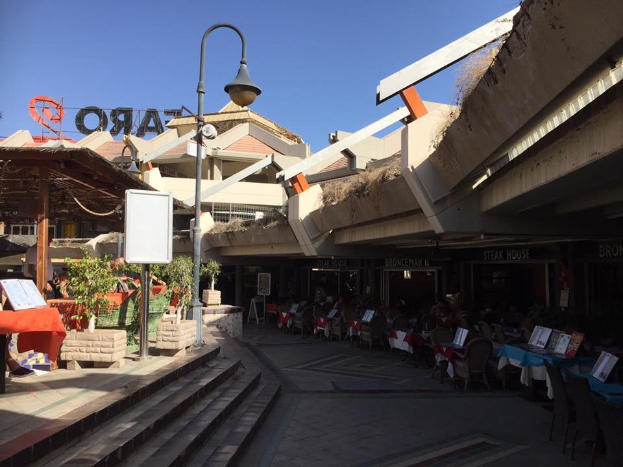 Eine Straße mit Tischen und Stühlen in der Mitte, umgeben von Gebäuden, Straßenmöbeln und Grün, unter einem klaren blauen Himmel, mit Büchern und Gegenständen auf den Tischen.