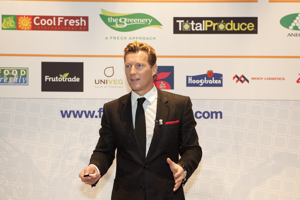 Ein Mann in einem schwarzen Blazer hält ein Objekt und spricht, mit einem weißen Banner und einer weißen Tafel mit Organisationslogos im Hintergrund.