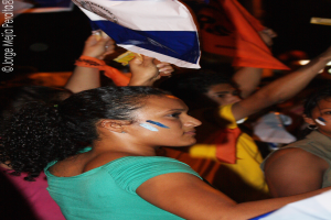 Eine Frau in einem grünen T-Shirt steht auf einer Demonstration und hölt ein weißes Schild in der Hand, mit vielen anderen Menschen, die hinter ihr stehen.
