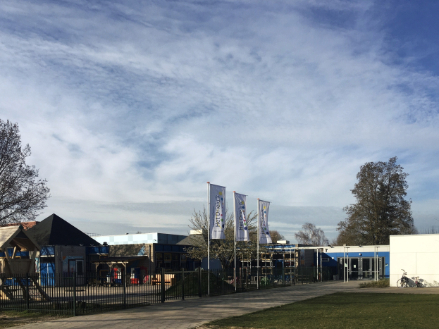 Großes Gebäude mit Metallzaun, Rasenfläche, Weg, Bäumen, Bannern, Fahrrad, nahen Häusern und bewölktem Himmel, identifiziert als neuer Spielplatz an der Schule.
