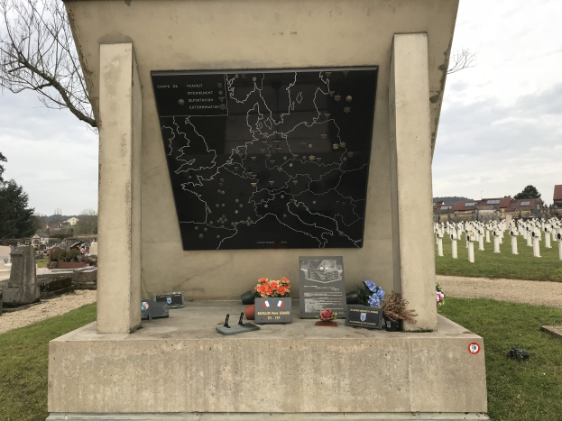 Ein Friedhof-Denkmal mit einer Karte, umgeben von Blumen und Gegenständen, das das Grab eines deutschen Weltkriegsoldaten markiert, mit Gräbern, Bäumen, Gebäuden und einem blauen Himmel im Hintergrund.