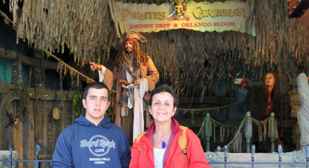 Ein Mann und eine Frau stehen vor einem Piratenschiff mit einem Geländer am Boden, Statuen von kostümierten Menschen, einem Banner mit Text und Lichtern im Hintergrund.