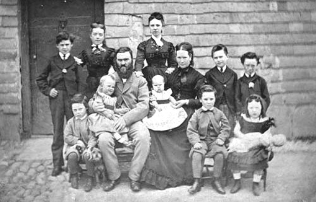 Schwarzes und weißes Foto einer Familie, die vor einem Haus posiert, wobei einige auf einer Bank sitzen und andere stehen, eine Wand und eine Tür im Hintergrund sichtbar.