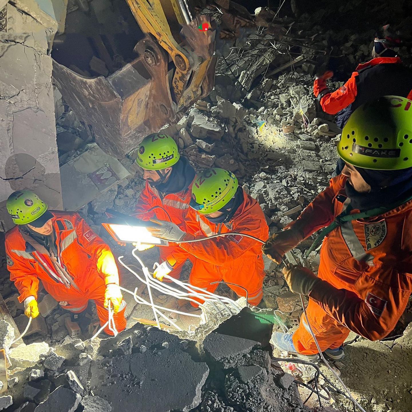 Eine Gruppe von Rettungskräften in orangefarbenen Uniformen und grünen Helmen beseitigt Trümmmer von einem eingestürzten Gebäude, wobei sie Werkzeuge und Materialien verwenden, mit einem Kran im Hintergrund.