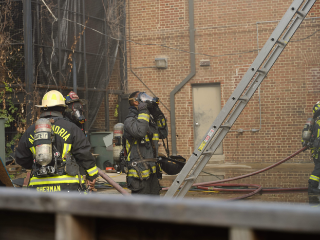 Eine Gruppe von Feuerwehrleuten in Helmen und Zylindern steht vor einem Gebäude mit einer Leiter, Rohren, Bäumen und anderen Gegenständen, mit einem Geländer am unteren Bildrand.