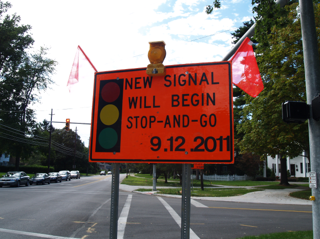 Eine Straße mit Fahrzeugen, eine Verkehrsampel mit einem Schild, das "Neues Signal Beginn Stopp und Gehen" anzeigt, Bäume, Gebäude und einen klaren blauen Himmel im Hintergrund.