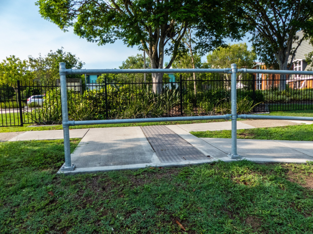 Metallzaun in einer grasigen Fläche mit umgebenen Pflanzen, Bäumen und einem Auto auf der Straße, mit Gebäuden und einem klaren blauen Himmel im Hintergrund, wahrscheinlich Teil eines Spielplatzes.