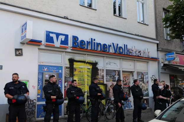 Eine Gruppe von Polizeibeamten steht vor einer Berliner Volksbank, einige halten Helme, mit einem Auto auf der rechten Seite, einem Baum auf der linken Seite und Gebäuden mit Fenstern und Texttafeln im Hintergrund.